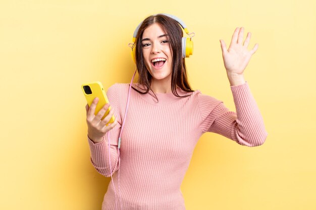 Giovane donna ispanica che sorride felicemente, agitando la mano, accogliendoti e salutandoti. concetto di cuffie e telefono