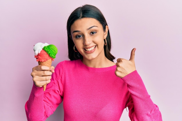 Giovane donna ispanica che tiene il gelato sorridendo felice e positivo, pollice in su facendo eccellente e segno di approvazione