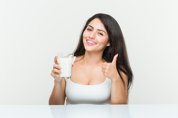 Giovane donna ispanica che tiene un bicchiere di latte che sorride e che solleva pollice su