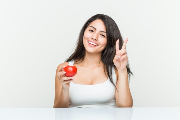 Giovane donna ispanica che tiene un pomodoro che mostra numero due con le dita.