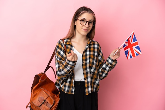 Giovane donna ispanica che tiene una bandiera del Regno Unito isolata sulla parete rosa che fa gesto di soldi