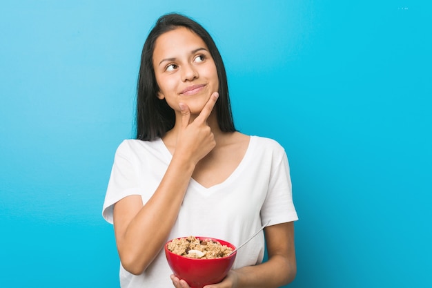 Giovane donna ispanica che tiene una ciotola di cereali guardando lateralmente con espressione dubbiosa e scettica.