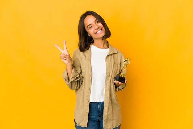 Giovane donna ispanica che tiene una statuetta del premio gioiosa e spensierata che mostra un simbolo di pace con le dita