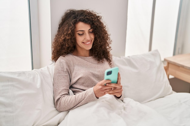 Giovane donna ispanica che usa lo smartphone seduto sul letto in camera da letto