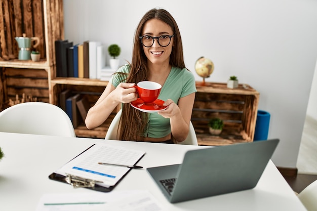 Giovane donna ispanica che utilizza il computer portatile che beve caffè seduto sul tavolo a casa