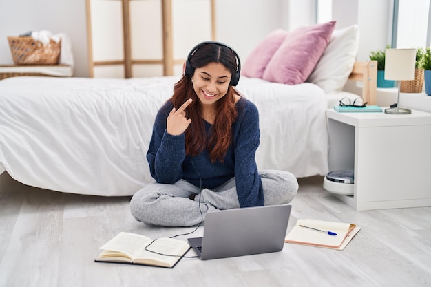 Giovane donna ispanica che utilizza il computer portatile che si siede sul pavimento alla camera da letto che sorride felice che indica con la mano e il dito