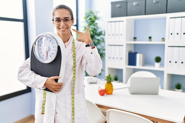 Giovane donna ispanica come medico nutrizionista che tiene la bilancia che punta il dito verso un auto sorridente felice e orgoglioso