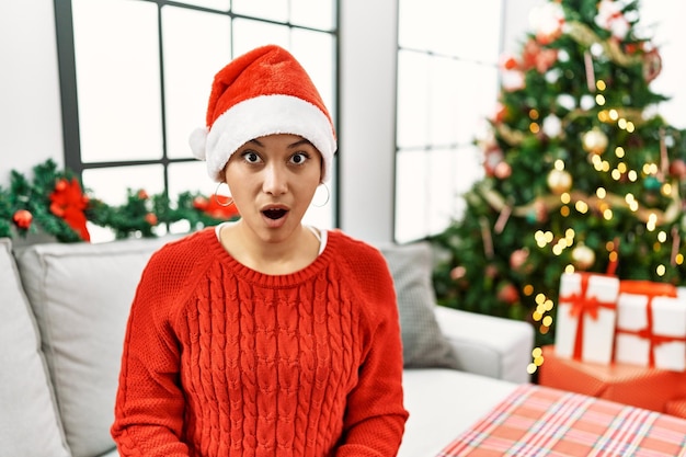 Giovane donna ispanica con i capelli corti che indossa un cappello di Natale seduto sul divano impaurito e scioccato dalla sorpresa e dall'espressione stupita paura e viso eccitato