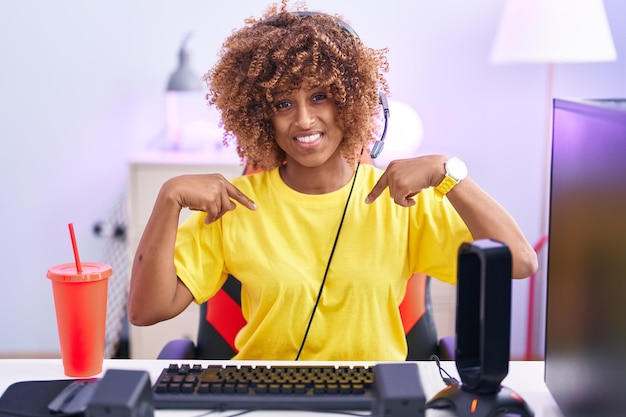 Giovane donna ispanica con i capelli ricci che gioca ai videogiochi con le cuffie che sembra sicura di sé con il sorriso sul viso che si indica con le dita orgogliose e felici