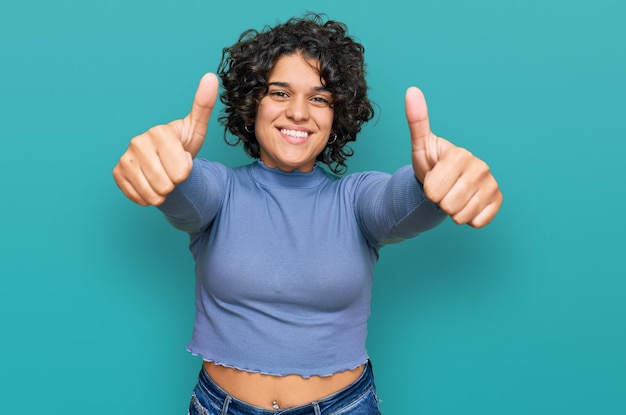 Giovane donna ispanica con i capelli ricci che indossa abiti casual che approva facendo un gesto positivo con il pollice in su sorridente e felice per il gesto del vincitore di successo