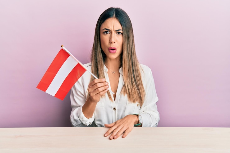 Foto giovane donna ispanica con la bandiera austriaca seduta sul tavolo spaventata e sbalordita con la bocca aperta per sorpresa faccia di incredulità