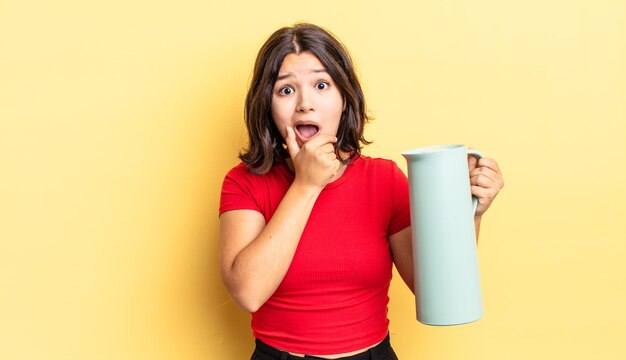 Giovane donna ispanica con la bocca e gli occhi spalancati e la mano sul mento. concetto di thermos