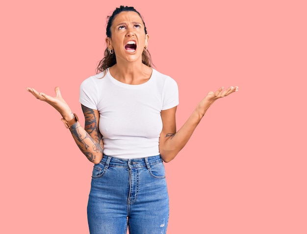 Giovane donna ispanica con tatuaggio che indossa una maglietta bianca casual pazza e pazza che grida e urla con espressione aggressiva e braccia alzate concetto di frustrazione