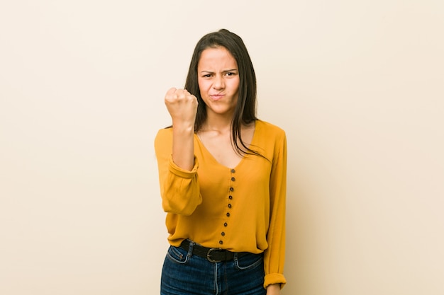 Giovane donna ispanica contro un muro beige che mostra il pugno con con espressione facciale aggressiva.