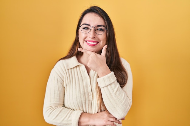 Giovane donna ispanica in piedi su sfondo giallo che guarda fiducioso alla telecamera sorridendo con le braccia incrociate e la mano alzata sul mento. pensare positivo.