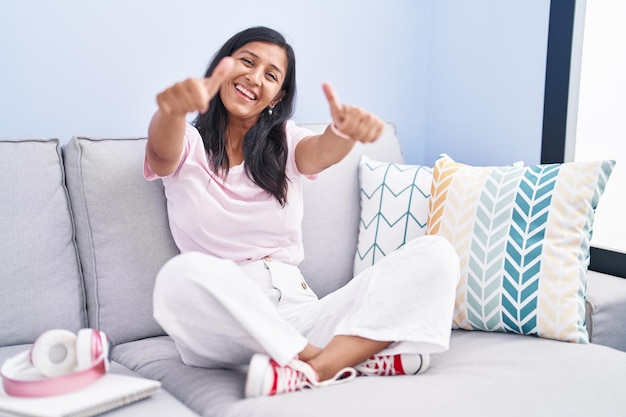 Giovane donna ispanica seduta sul divano di casa che approva facendo un gesto positivo con il pollice in alto sorridente e felice per il gesto del vincitore di successo