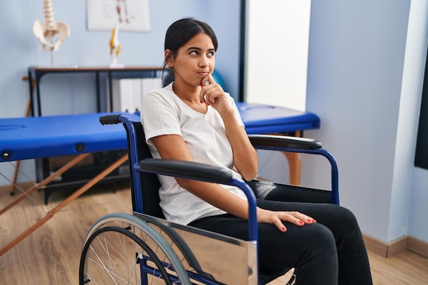 Giovane donna ispanica seduta sulla sedia a rotelle presso la clinica di fisioterapia pensando concentrata sul dubbio con il dito sul mento e alzando lo sguardo chiedendosi