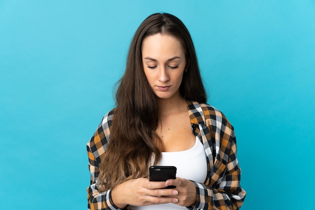 Giovane donna ispanica sopra la parete blu isolata utilizzando il telefono cellulare