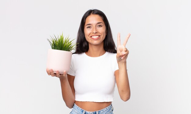 giovane donna ispanica sorridente e dall'aspetto amichevole, mostrando il numero due e tenendo in mano una pianta decorativa della casa
