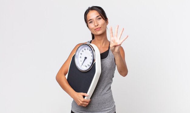 Giovane donna ispanica sorridente e dall'aspetto amichevole, mostrando il numero quattro e tenendo in mano una bilancia. concetto di dieta