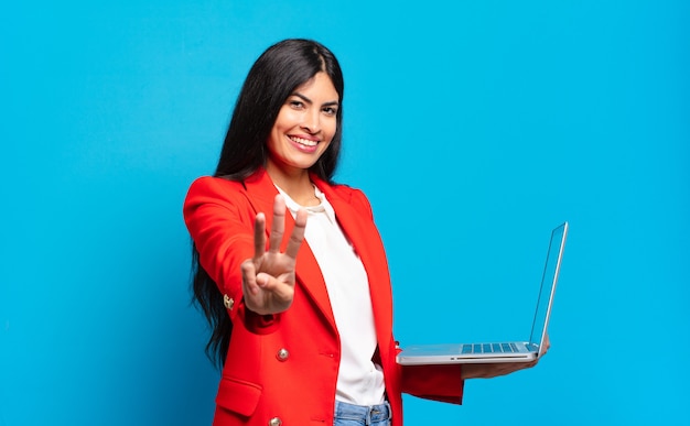 Giovane donna ispanica sorridente e dall'aspetto amichevole, mostrando il numero tre o terzo con la mano in avanti, il conto alla rovescia. concetto di laptop