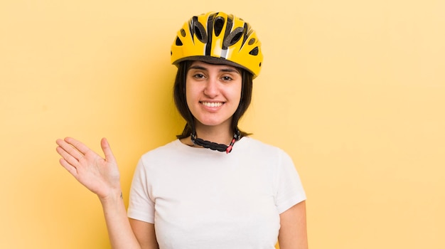 Giovane donna ispanica sorridente felicemente agitando la mano dandoti il benvenuto e salutandoti concetto di casco da bici