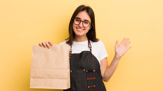 Giovane donna ispanica sorridente felicemente agitando la mano dandoti il benvenuto e salutandoti il concetto di consegna di fast food