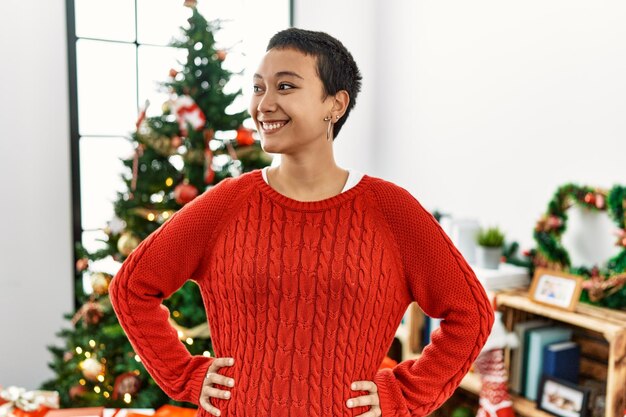 Giovane donna ispanica sorridente fiducioso in piedi accanto all'albero di Natale a casa