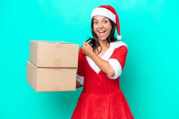 Giovane donna ispanica vestita da mama noel che tiene una scatola di cartoni animati isolata su sfondo blu che celebra una vittoria