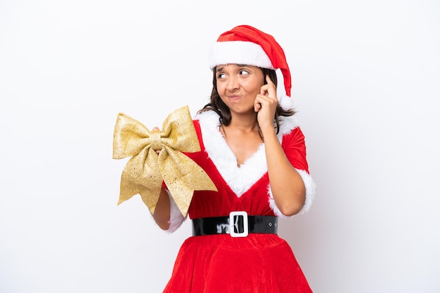 Giovane donna ispanica vestita da mama noel in possesso di un fiocco di Natale isolato su sfondo bianco frustrato e che copre le orecchie