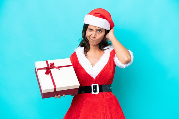 Giovane donna ispanica vestita da mama noel in possesso di un regalo isolato su sfondo blu con dubbi