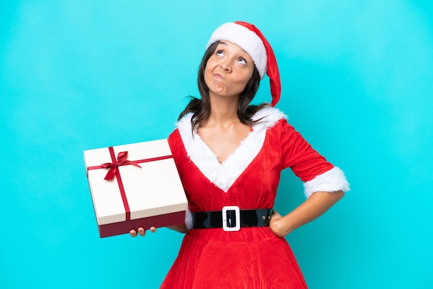 Giovane donna ispanica vestita da mama noel in possesso di un regalo isolato su sfondo blu e alzando lo sguardo