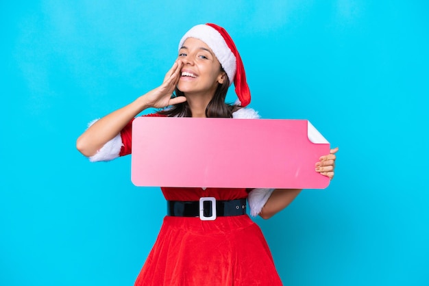 Giovane donna ispanica vestita da mama noel isolata su sfondo blu con in mano un cartello vuoto e gridando