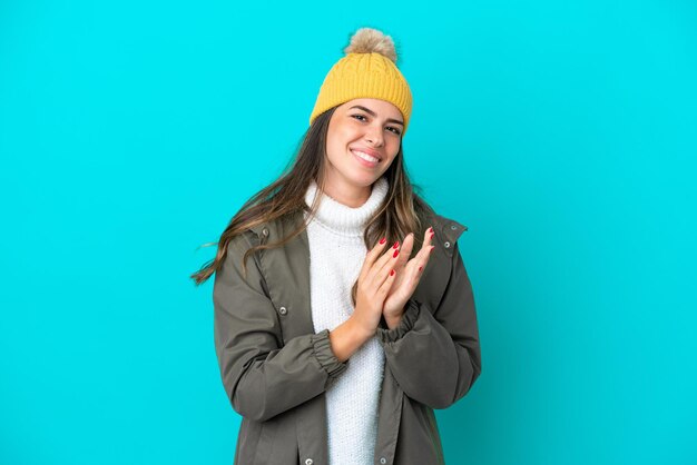 Giovane donna italiana che indossa giacca invernale e cappello isolati su sfondo blu che applaude dopo la presentazione in una conferenza