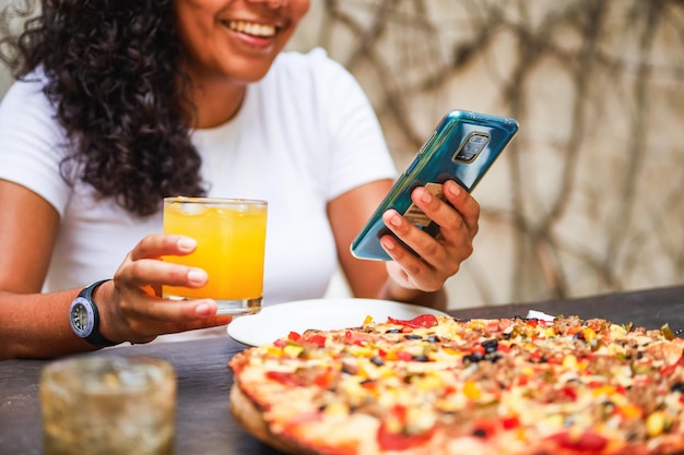 Giovane donna latina al ristorante usando il suo smartphone con una bevanda in mano pizza sul tavolo