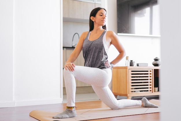 Giovane donna latina che allunga i muscoli flessori dell'anca su un tappetino da yoga nel suo salotto