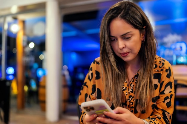 Giovane donna latina che guarda il dispositivo mobile all'interno nel ritratto di un ristorante o di un pub