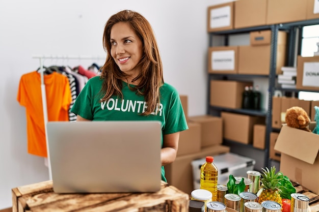 Giovane donna latina che indossa l'uniforme volontaria utilizzando il computer portatile che lavora al centro di beneficenza