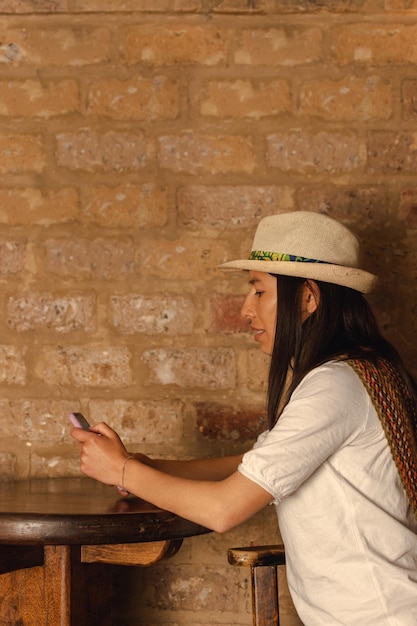Giovane donna latina che indossa un cappello seduto in un ristorante guardando il suo telefono cellulare contro il muro di fango