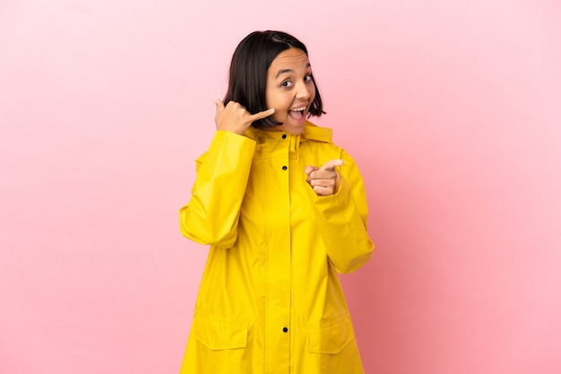 Giovane donna latina che indossa un cappotto antipioggia su sfondo isolato che fa il gesto del telefono e indica la parte anteriore