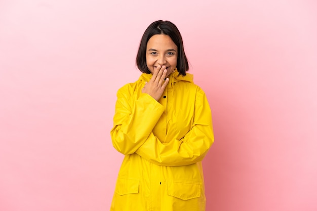 Giovane donna latina che indossa un cappotto antipioggia su un muro isolato felice e sorridente che copre la bocca con la mano