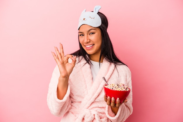 Giovane donna latina che indossa un pigiama in possesso di una ciotola di cereali isolata su sfondo rosa allegro e fiducioso che mostra gesto ok.