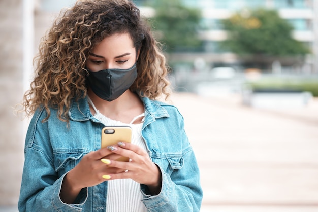 Giovane donna latina che indossa una maschera facciale mentre usa il suo telefono cellulare all'aperto in strada. Nuovo stile di vita normale. Concetto urbano.