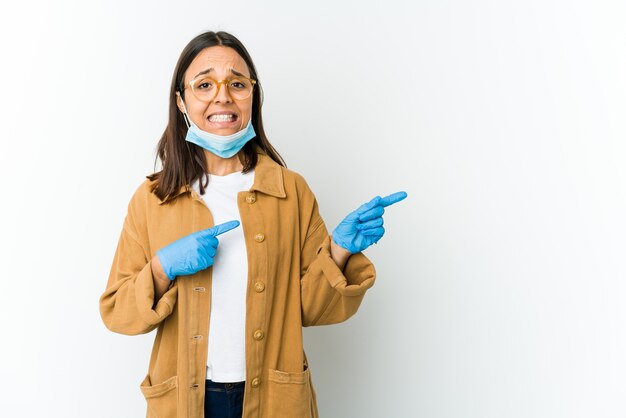 Giovane donna latina che indossa una maschera per proteggere da covid isolato sul muro bianco scioccato che punta con il dito indice a una copia spazio