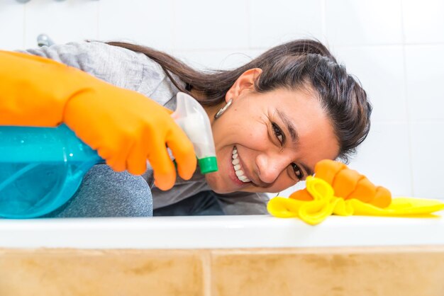 Giovane donna latina che pulisce la vasca da bagno sorridente che dà una occhiata attraverso i prodotti per la pulizia