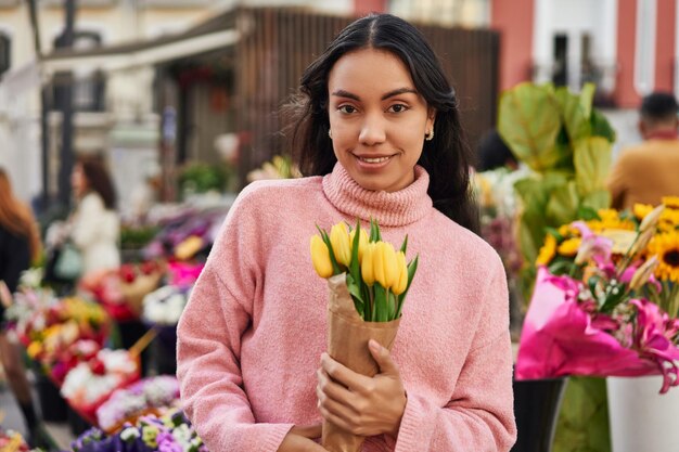 Giovane donna latina che si gode il profumo dei tulipani gialli mentre li acquista dalla bancarella di un venditore di piante