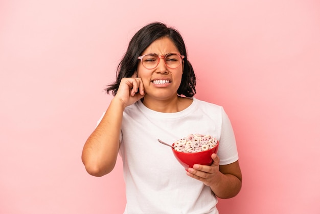 Giovane donna latina che tiene i cereali isolati su sfondo rosa che copre le orecchie con le mani.