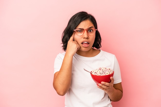 Giovane donna latina che tiene i cereali isolati su sfondo rosa che mostra un gesto di delusione con l'indice.