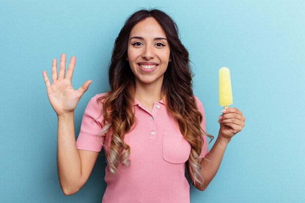 Giovane donna latina che tiene il gelato isolato su sfondo blu sorridente allegro che mostra il numero cinque con le dita.