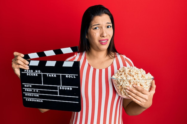 Giovane donna latina che tiene l'assicella del film video e il concetto di dubbio di espressione all'oscuro e confuso del popcorn